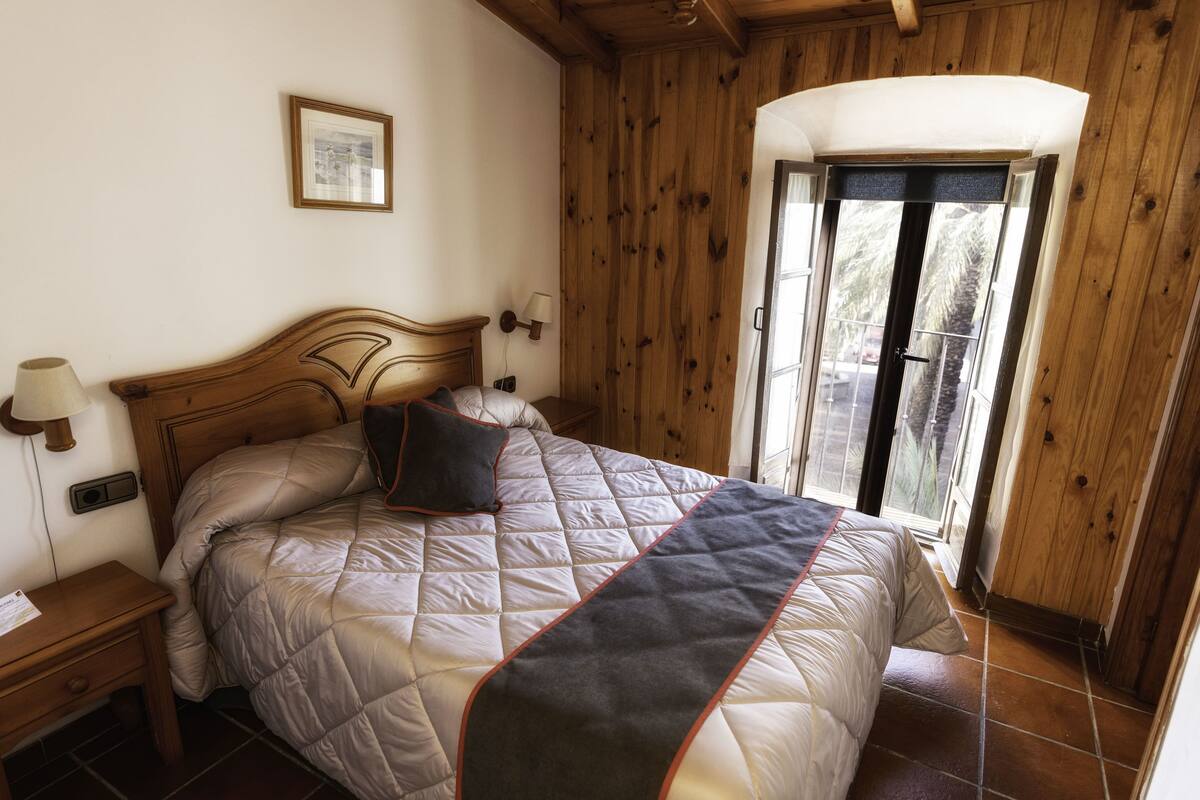 Un dormitorio con paneles de madera, una cama, una mesita de noche, una lámpara de pared y una ventana con vistas al exterior.