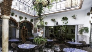 Un patio con mesas redondas y sillas de mimbre, rodeado de plantas en macetas y una fuente decorativa.