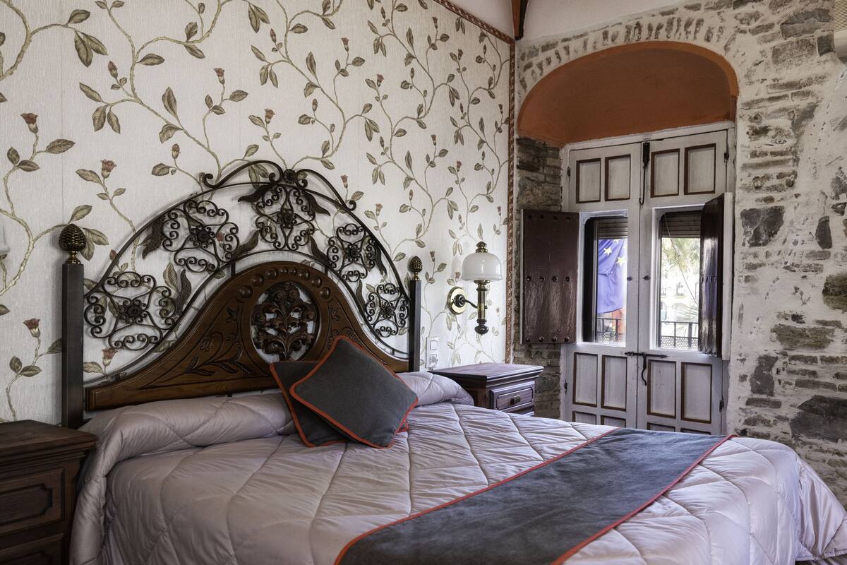 Un dormitorio con una cama grande, un cabecero decorativo, una mesita de noche, una lámpara de pared y una ventana con contraventanas.