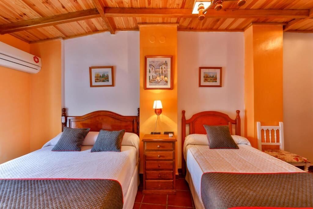 A hotel room with two beds, wooden headboards, a nightstand with a lamp, and a wall-mounted air conditioner.