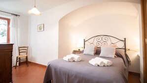 Un letto rifatto con cuscini e asciugamani arrotolati, una sedia di legno, un comodino con una lampada e una finestra con tende.
