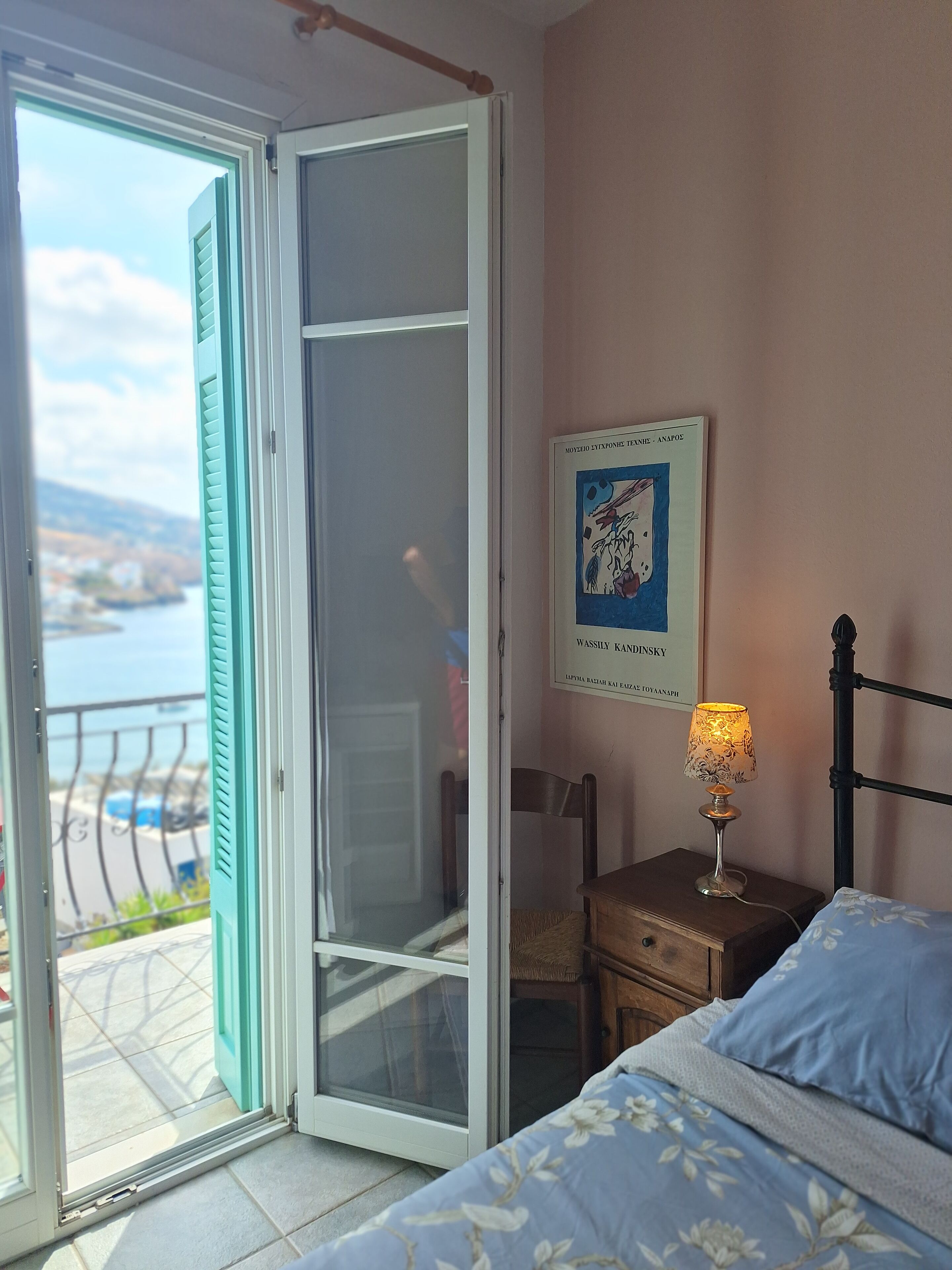 Un dormitorio con cama, mesita de noche, lámpara, una silla, una puerta y vistas al exterior.