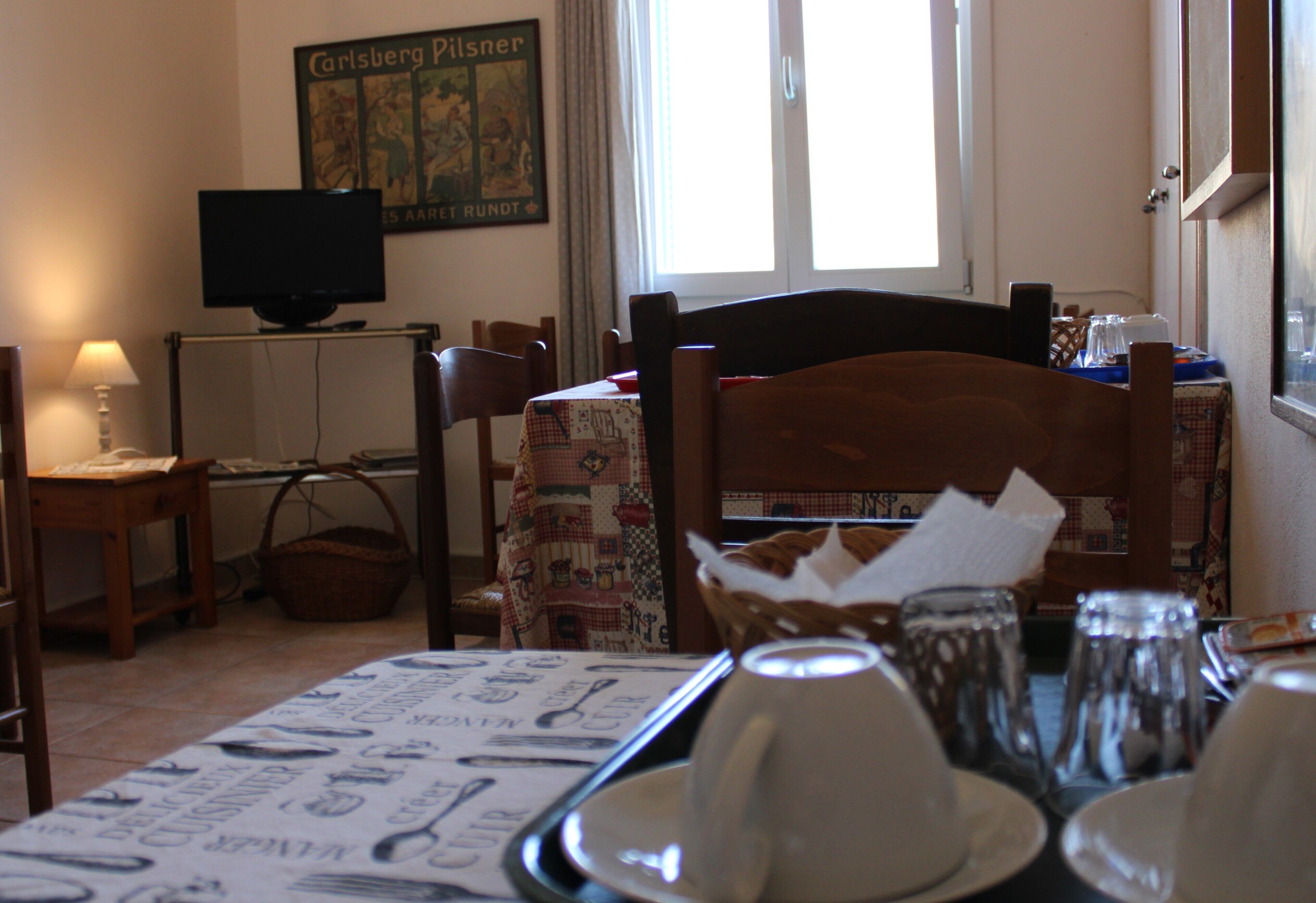 Un coin repas avec une table dressée pour deux personnes, un téléviseur posé sur un meuble et une fenêtre avec des rideaux.
