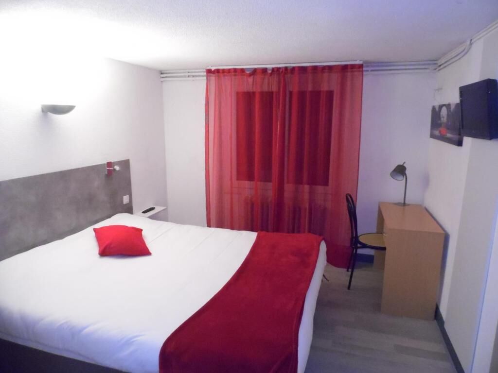 A hotel room with a bed, a desk, a chair, a red curtain, and a picture on the wall.