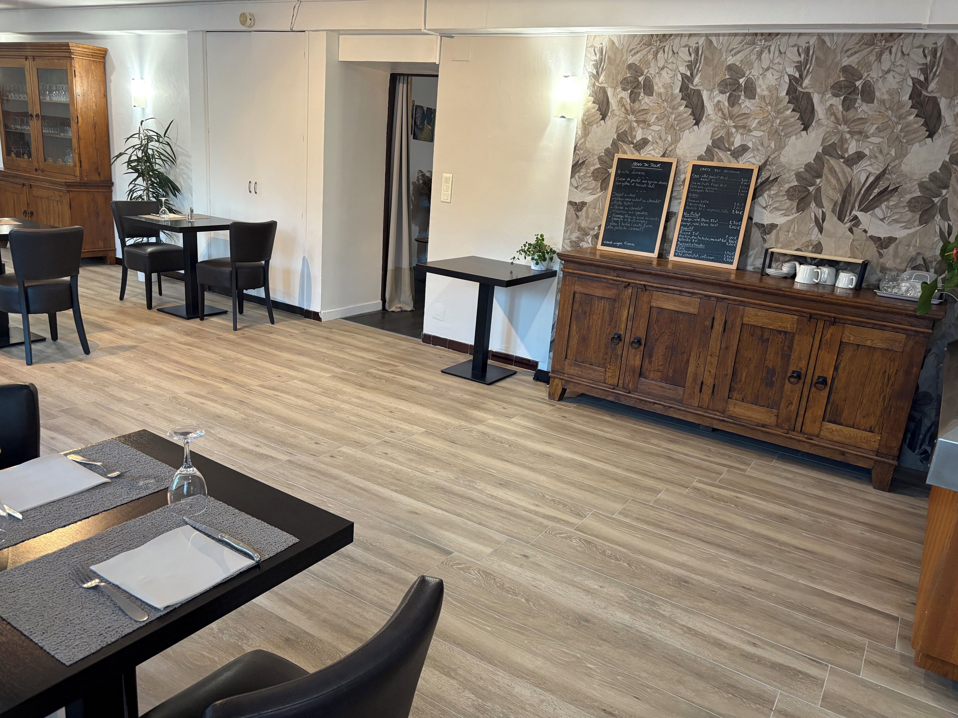 A modern dining area with wooden flooring, black dining tables, and chairs. There is a wooden cabinet with glass doors and a menu board on the wall.