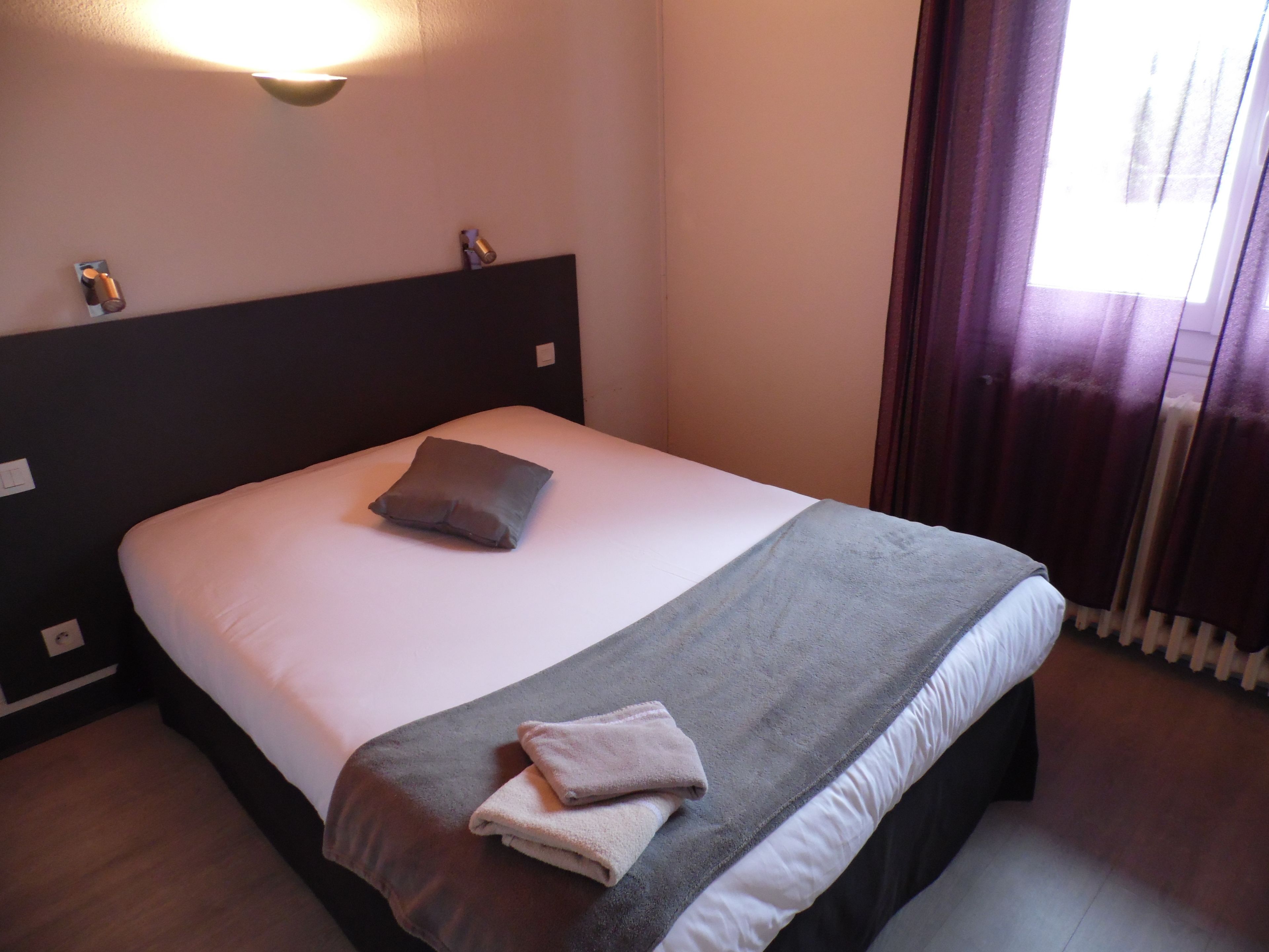Un lit bien fait, avec un oreiller et deux serviettes pliées, un luminaire fixé au mur et une fenêtre avec des rideaux.