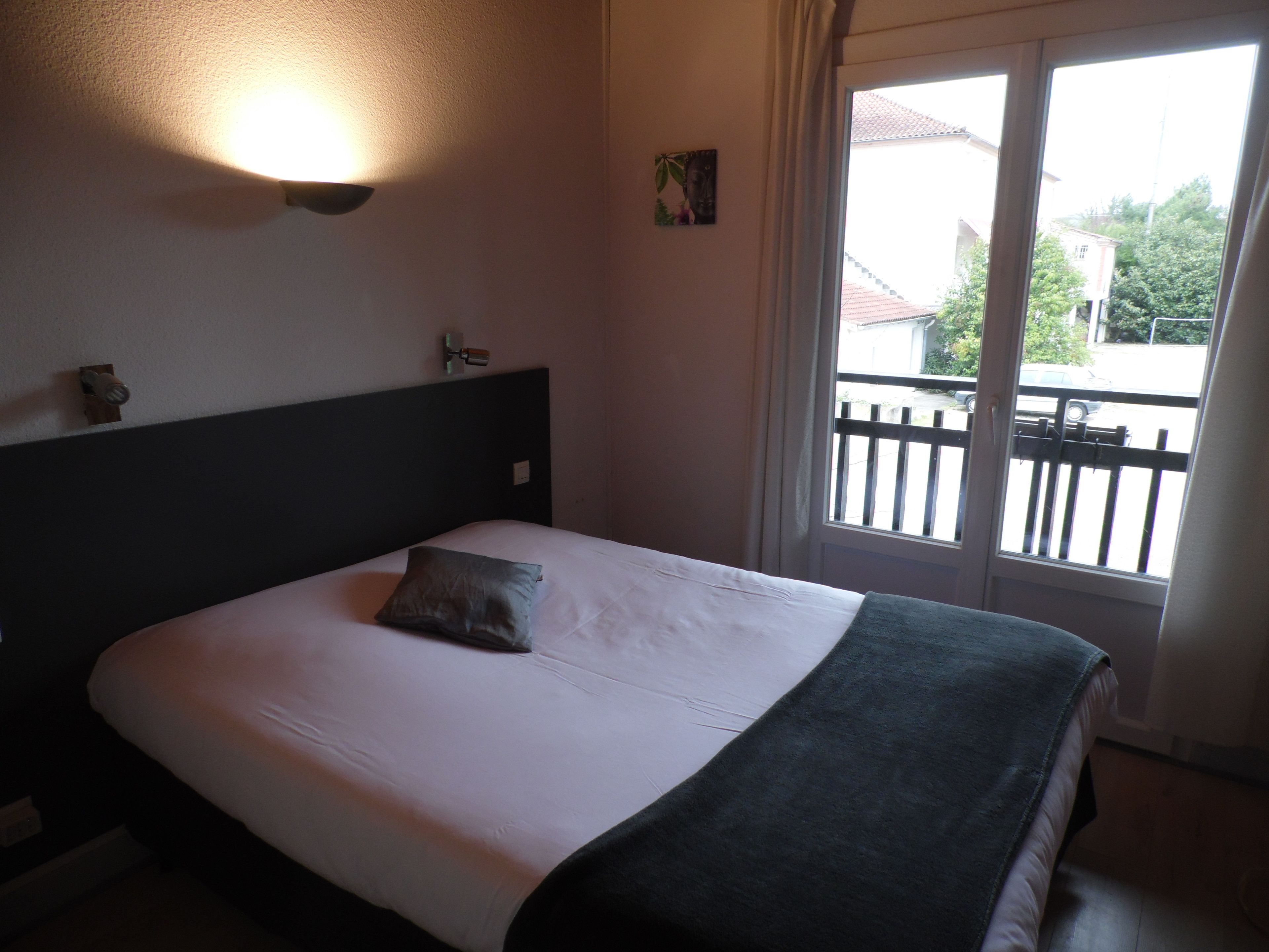 Quarto de hotel com cama, mesa de cabeceira, luminária na parede, janela com vista exterior e porta.