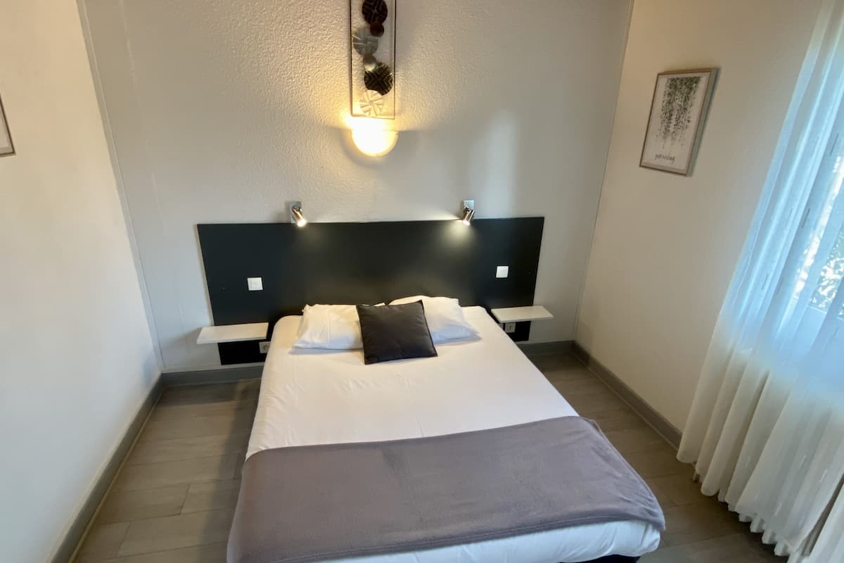 Een net opgemaakt bed met een wit-grijze dekbedovertrek, een zwart kussen en een nachtkastje met een lamp.
