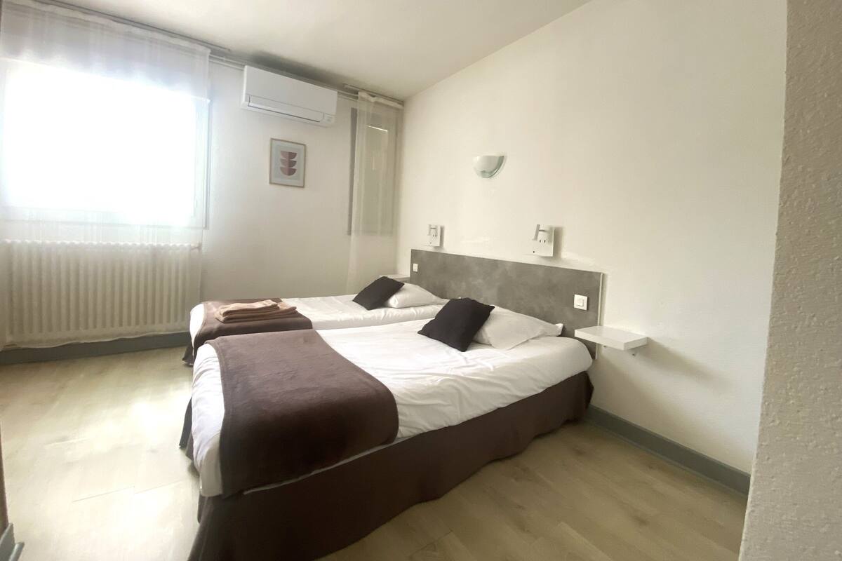 Een hotelkamer met twee bedden, een radiator, een airconditioner en een raam met dunne gordijnen.