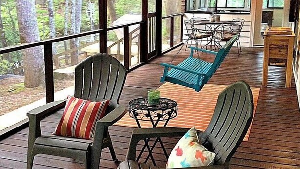 Eine überdachte Veranda mit Adirondack-Stühlen, einem Tisch und Blick auf einen Wald.
