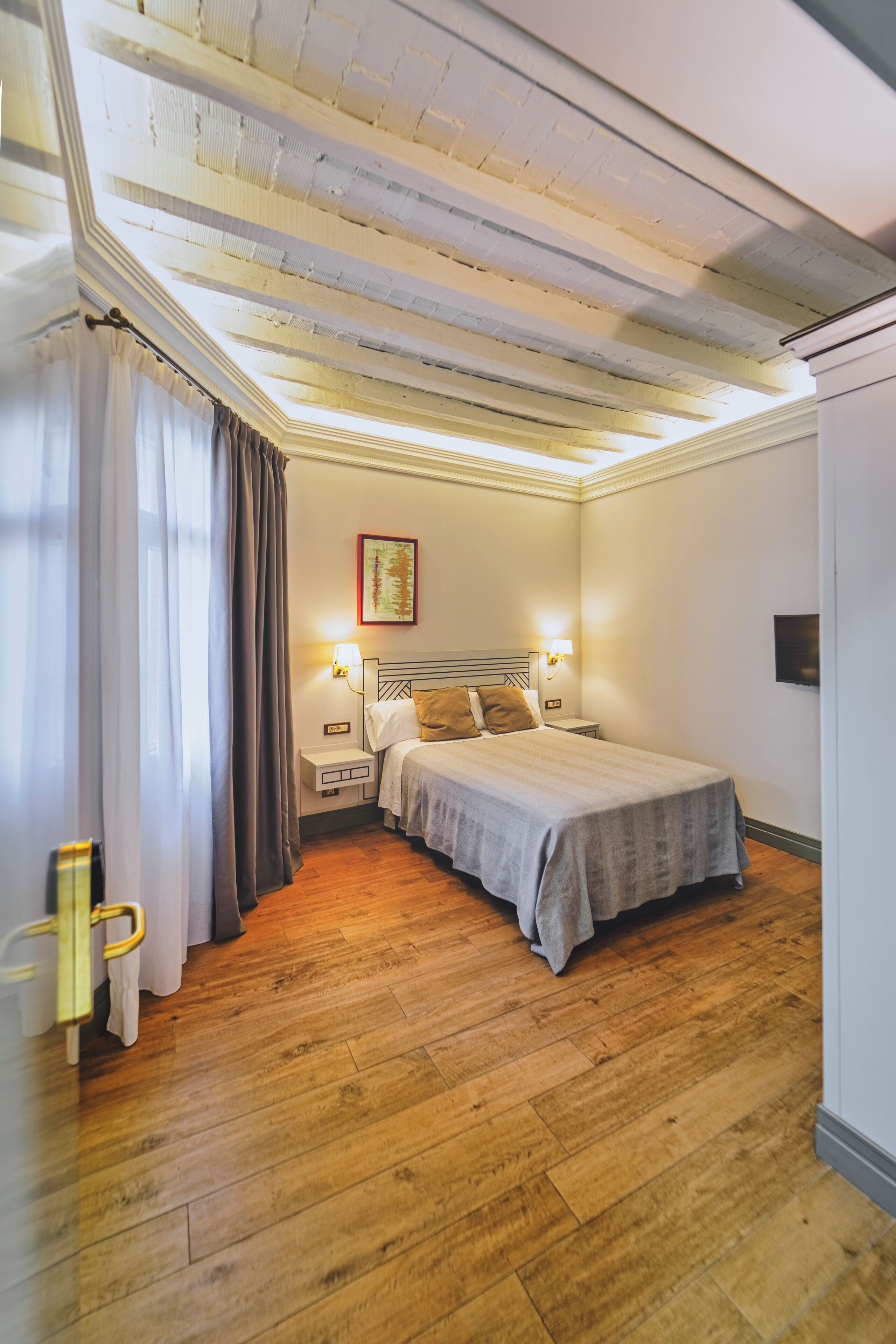 Un dormitorio con piso de madera, una cama con cabecera, una mesita de noche, una lámpara de pared y un ventanal con cortinas.