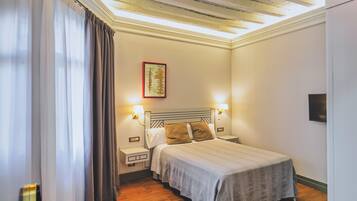 A bedroom with a wooden floor, a bed with a headboard, a nightstand, a wall-mounted lamp, and a window with curtains.