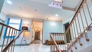 Una escalera con barandilla de madera y balaustres de metal, que conduce a un rellano con una lámpara y vista a otra habitación.
