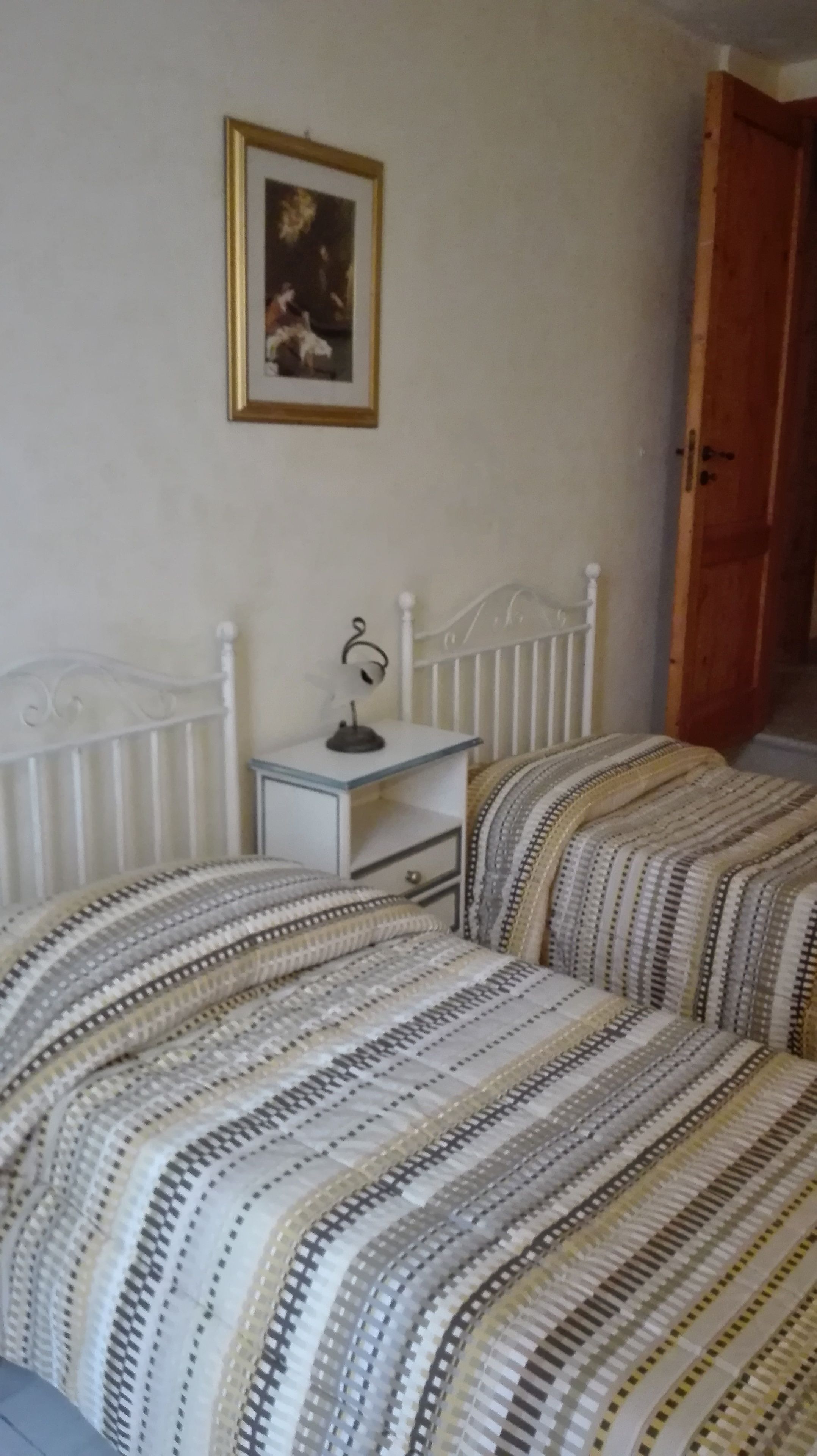 A bedroom with two beds, a bedside table with a lamp, a framed picture on the wall, and a wooden door.
