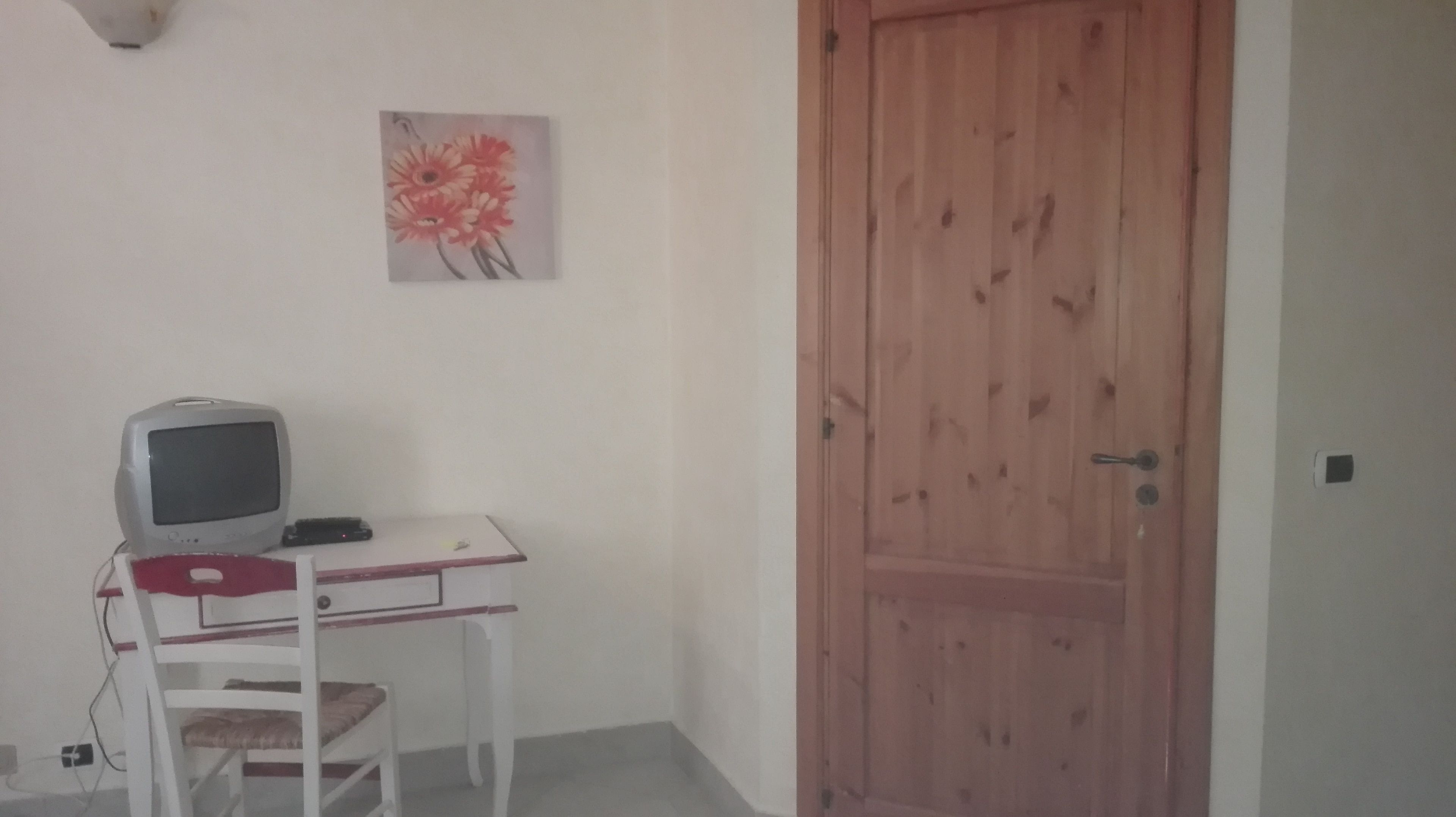 Ein Zimmer mit Holztür, Schreibtisch, Computer und Stuhl sowie einem Bild an der Wand.