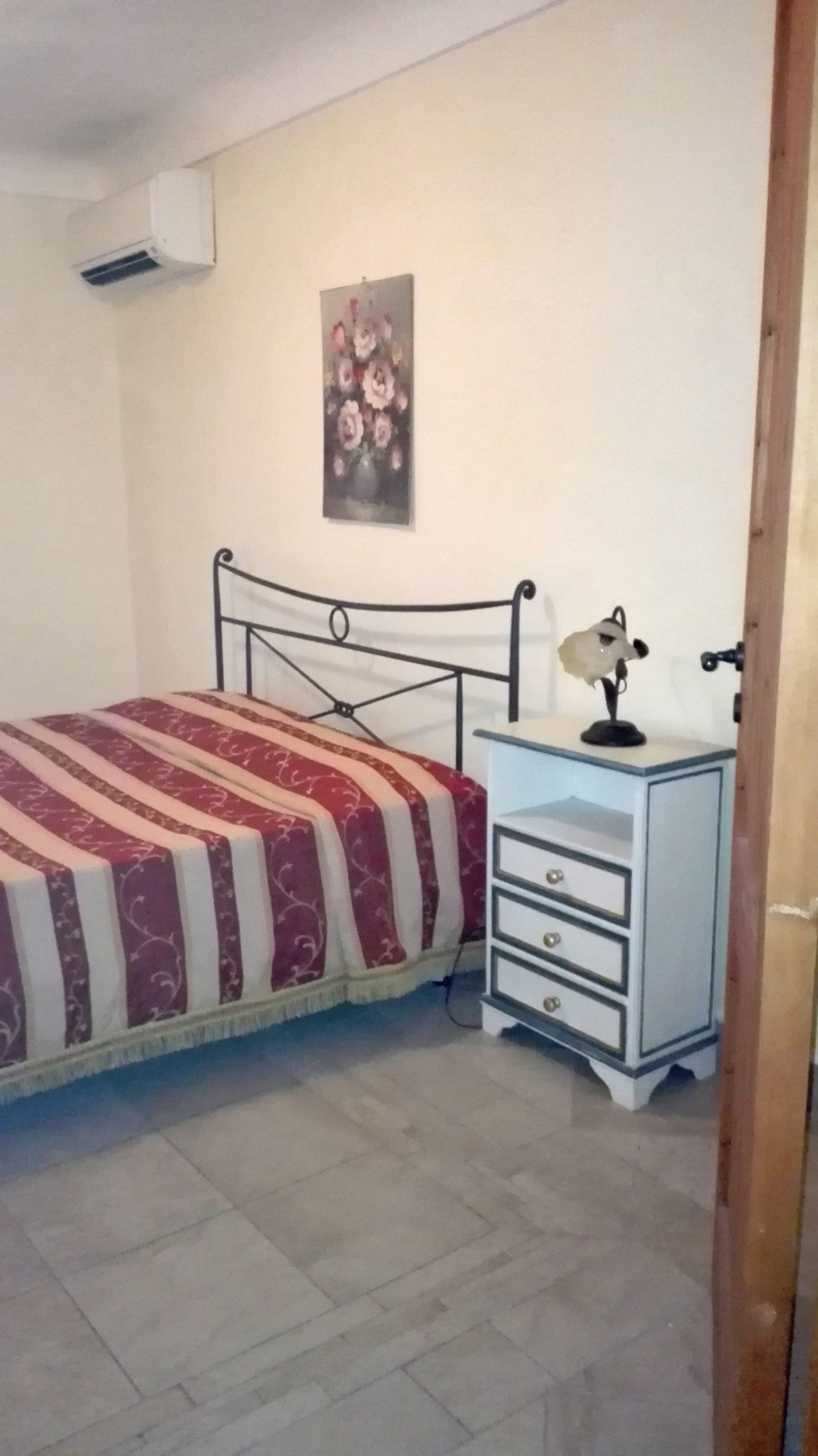 A bedroom with a bed, a nightstand, a lamp, and a wall-mounted air conditioner.