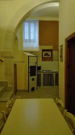 A dining area with a high table and chairs, a kitchen with a stove and cabinets, and a window with curtains. - B&B Candido Vacanze (Modica)