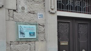 Fachada de edificio con pared de piedra, una placa con número y una puerta de madera oscura con ornamentación metálica.