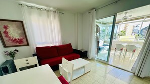 Un salon moderne avec un canapé rouge, une table basse blanche et une table à manger. Une porte-fenêtre coulissante donne accès à un espace extérieur agrémenté de chaises blanches.