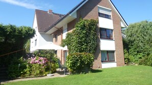A two-story house with a brick facade, a tiled roof, and a garden with pink flowers and a chair. - Apartment 5, 2- room, 50 m², mezzanine floor, garden use - guest house Müller (Scharbeutz)