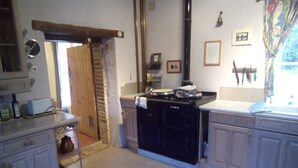 Een keuken met een fornuis, spoelbak en kastjes. Een houten deur leidt naar een andere kamer.
