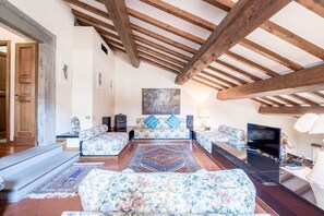 A spacious living room with a high ceiling, wooden beams, floral-patterned sofas, a large rug, and a television.