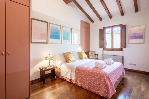 A bedroom with a bed, wooden floor, a nightstand, a lamp, a chair, a wardrobe, and a window.