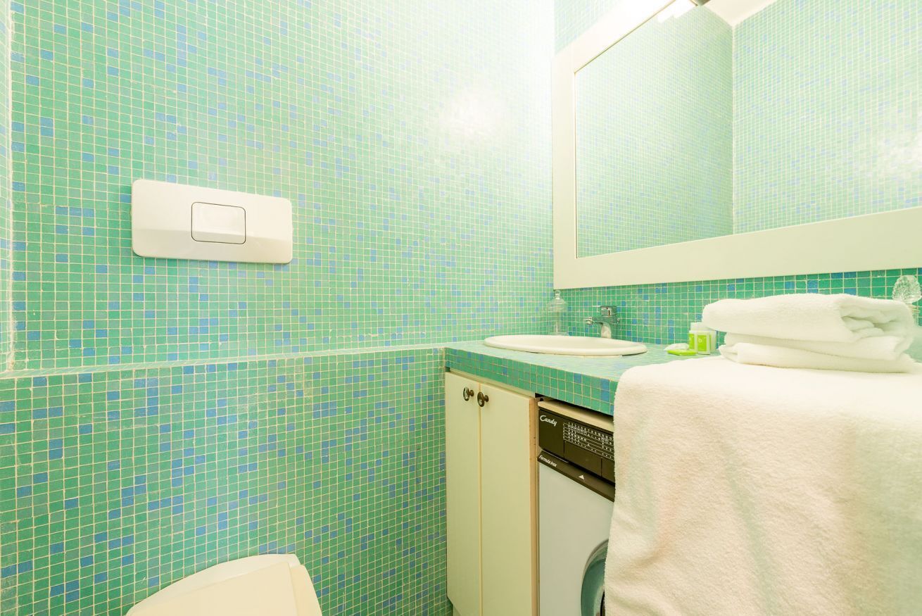 A bathroom with a green mosaic tile wall, a white sink, a mirror, a towel, and a dishwasher.