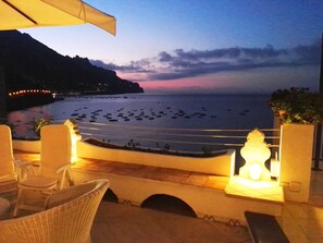 Terraço com cadeiras de vime e uma mesa, com vista para uma praia com barcos e um pôr do sol.