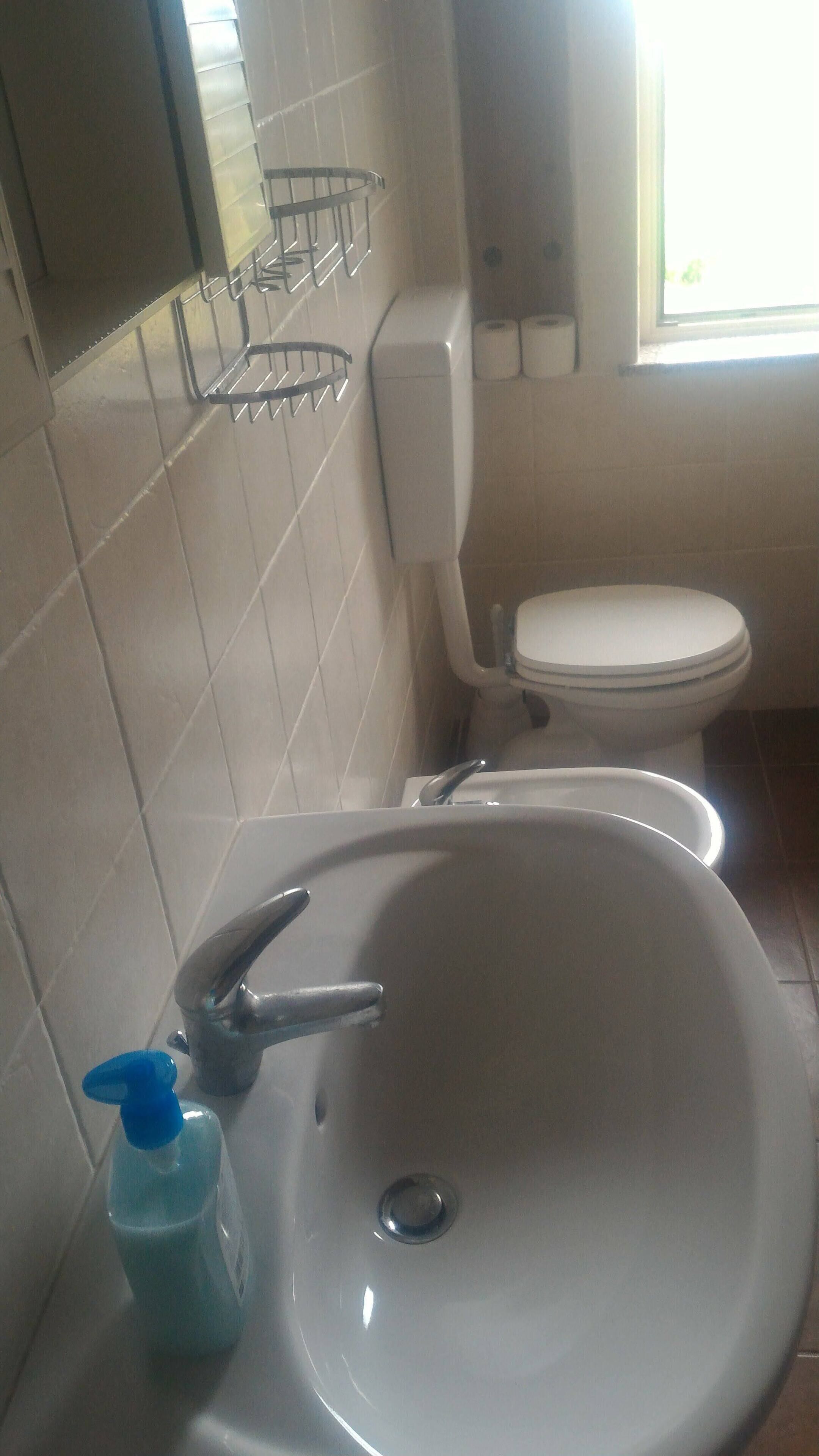 Een badkamer met een wit wastafel, een kraan, een zeepdispenser, een toilet en een raam.