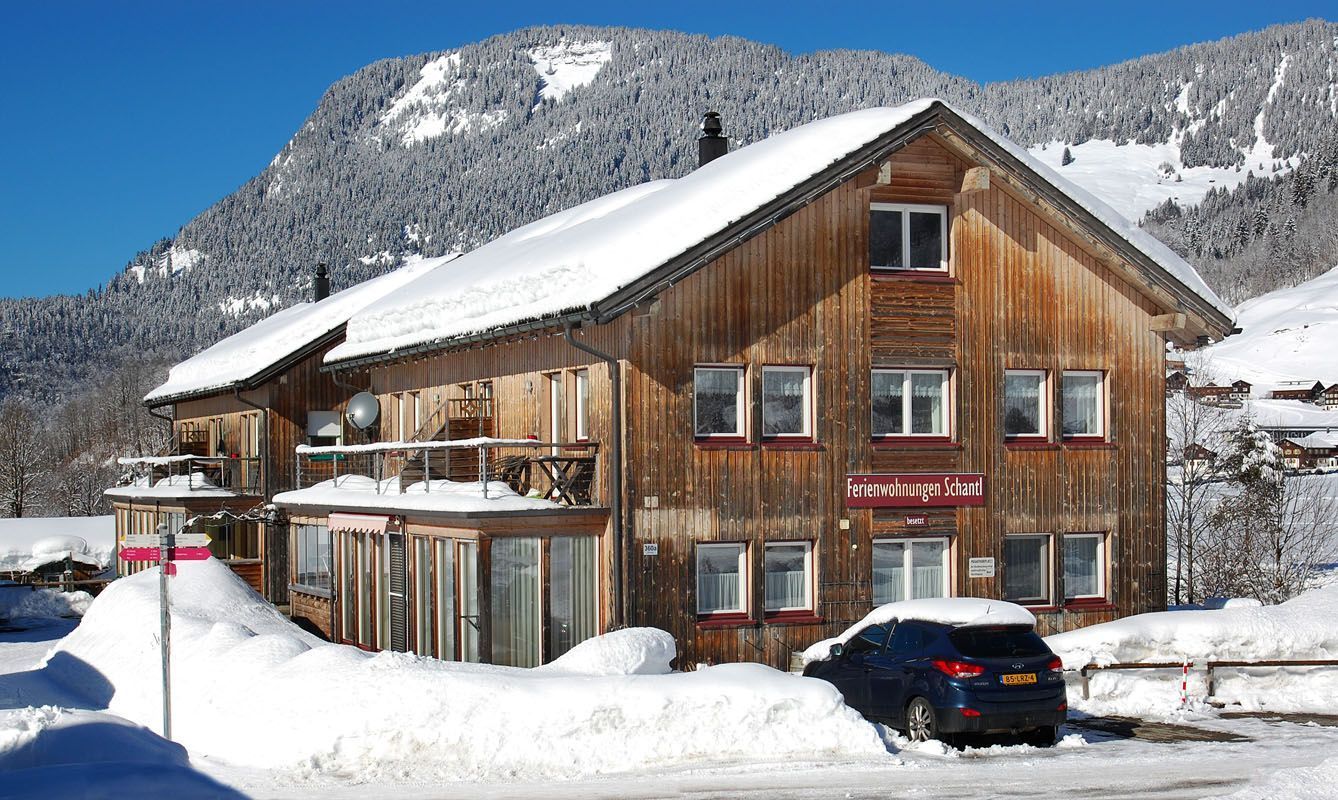 Een houten chalet met een balkon, een auto ervoor geparkeerd, en een bord waarop 'Ferienwohnungen Schand' staat.