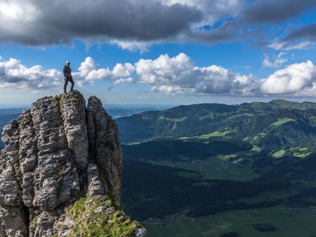 Eine Person steht auf einer felsigen Klippe mit Blick auf ein Tal und Berge im Hintergrund.