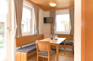 A room with a wooden bench, a table, a TV mounted on the wall, and a window with curtains.