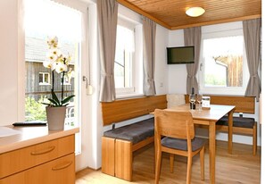 A room with a wooden bench, a table, a TV mounted on the wall, and a window with curtains.