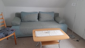 A small room with a grey sofa, a wooden chair, and a small table with a white tablecloth. - Sunset apartment (Fuhlendorf)