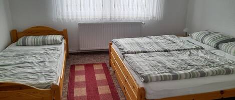 Una habitación con dos camas de madera, una ventana con cortinas translúcidas, un radiador y una alfombra de cuadros rojos y beige.