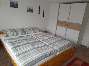 A single bed with striped bedding, a wooden headboard, a nightstand with a lamp, and a wardrobe with a wooden panel design. - Sunset apartment (Fuhlendorf)