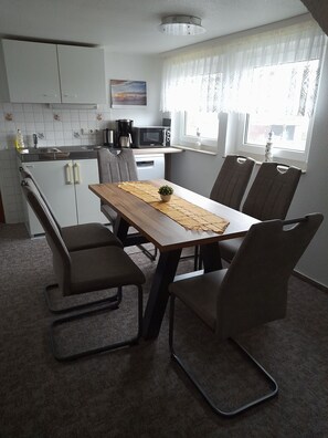 A compact kitchen with white cabinets, a microwave, and a coffee maker. There is a dining table with four chairs and a small potted plant. - Sunset apartment (Fuhlendorf)