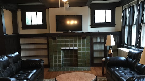 Een woonkamer met twee zwarte leren banken, een ronde salontafel, een open haard met groene tegels, een plafondventilator en een televisie boven de open haard.