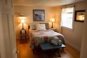 A cozy bedroom with a bed, two bedside tables with lamps, a framed picture on the left table, a window with curtains, and a bench with rolled towels.