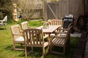 A wooden table with four chairs, a vase with flowers, and a grill in a backyard setting.