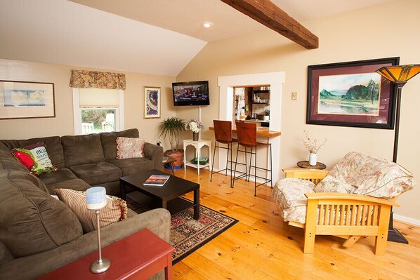A cozy living room with a brown sofa, a wooden coffee table, and a floral armchair. There is a TV mounted on the wall, a small dining area with a bar stool, and a framed painting on the wall.