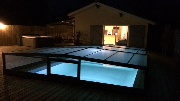 Una piscina con piso de vidrio, cubierta de madera y una casa al fondo.