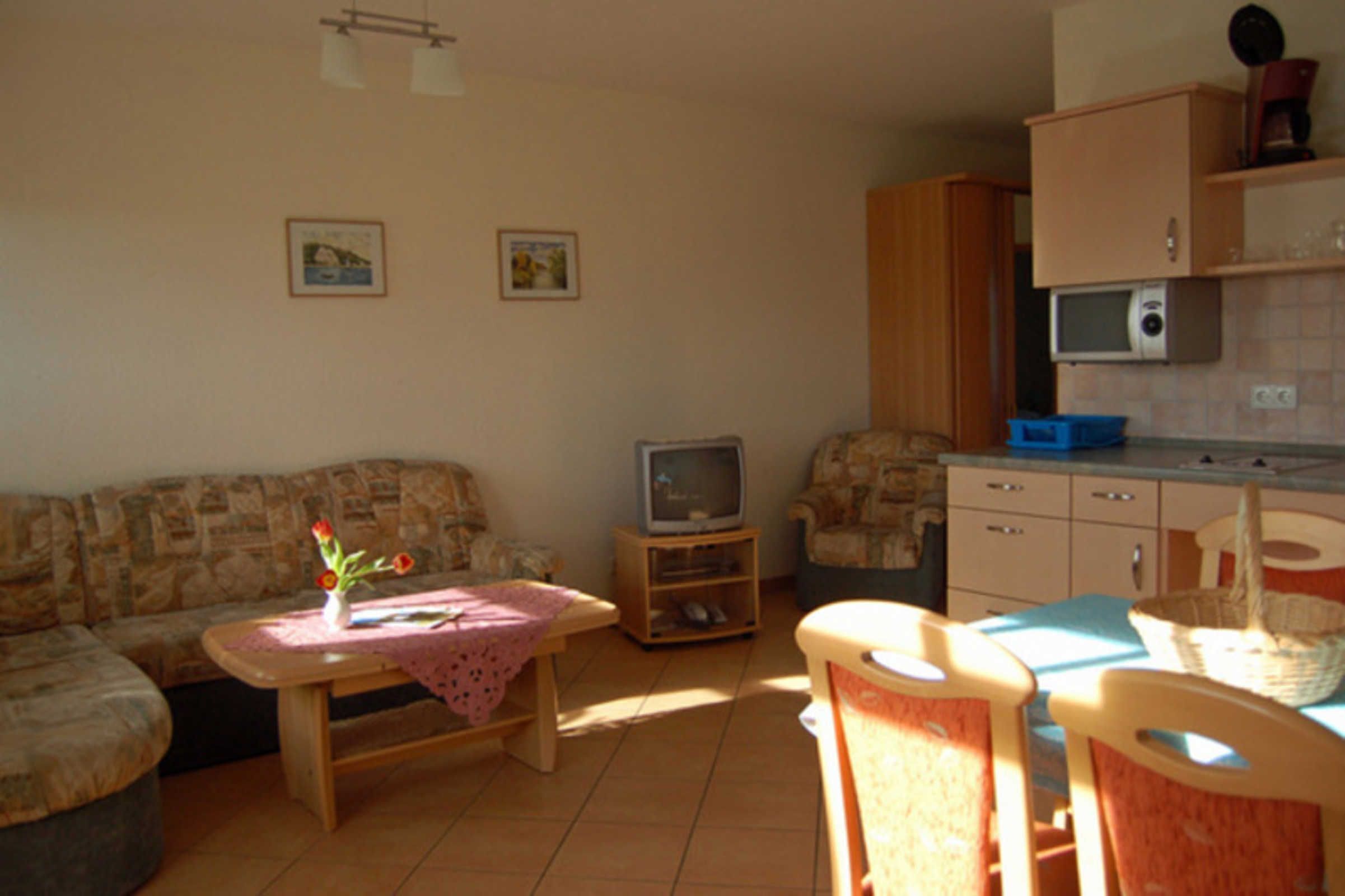 A living room with a sofa, a small table with a vase, a television, a kitchen area with cabinets and a microwave, and a dining table with chairs.