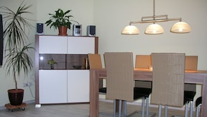 A dining area with a wooden table, wicker chairs, a potted plant, a cabinet with speakers, and a pendant light fixture. - Domizil Neu Venedig (Berlin)