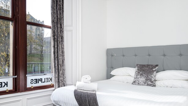 A hotel room with a bed, a window with curtains, and a view of a building outside. - Kelpies Serviced Apartments (Falkirk)