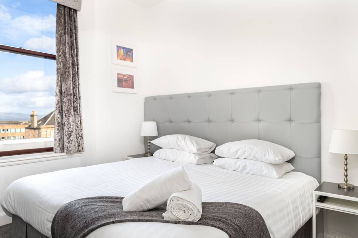 A hotel room with a large bed, a grey headboard, white bedding, a window with curtains, and a nightstand with a lamp.