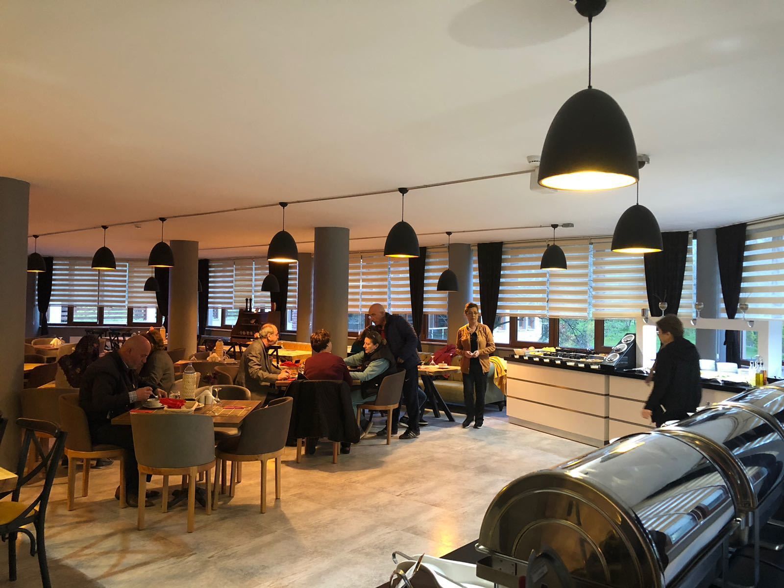 A modern dining area with multiple tables and chairs, large windows with blinds, and pendant lighting.