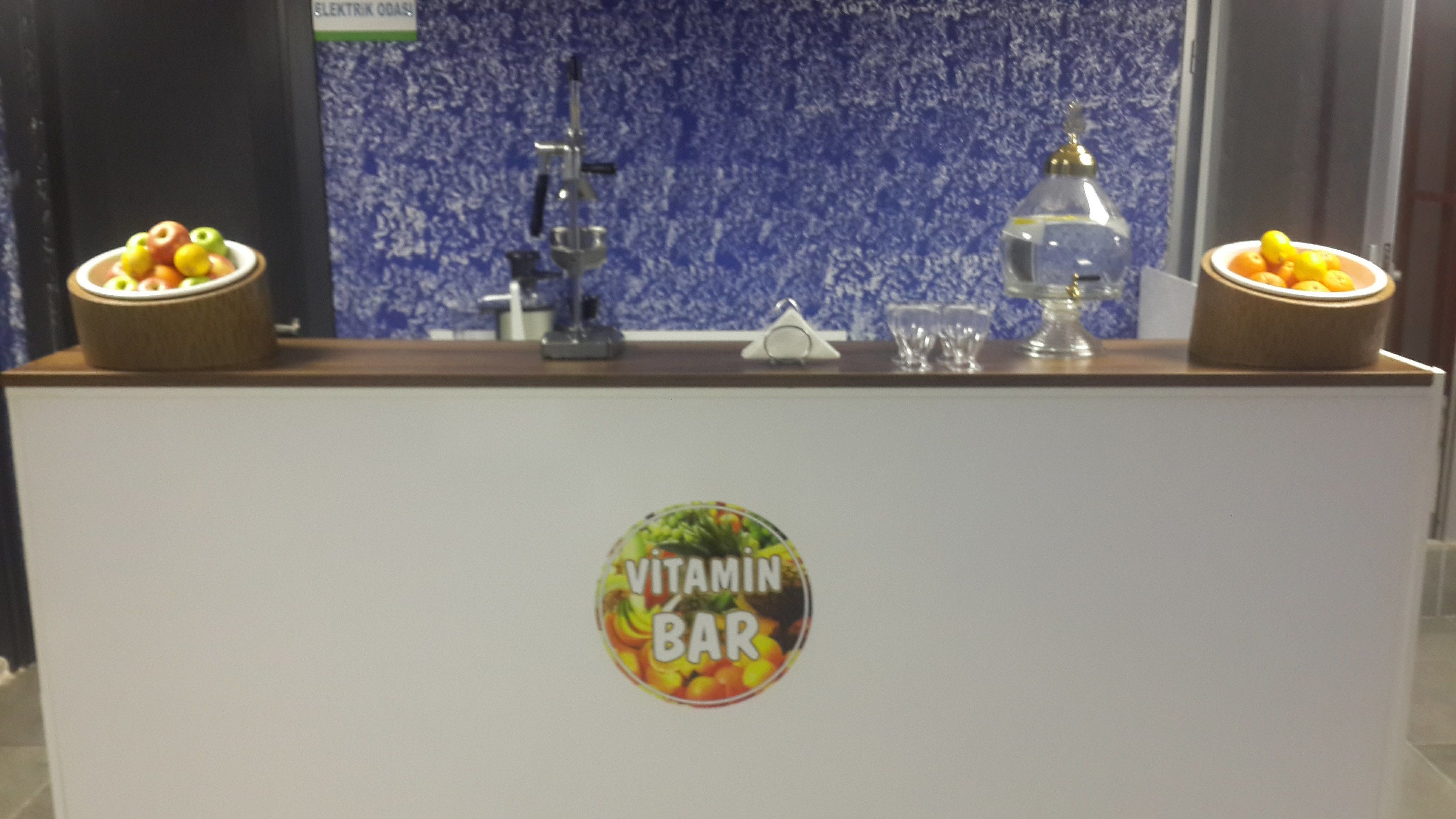 A counter with a 'Vitamin Bar' sign, two bowls of fruit, a water dispenser, and a glass.