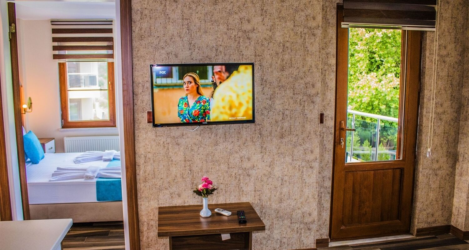 A hotel room with a television mounted on the wall, a wooden door, and a window with a view of greenery.